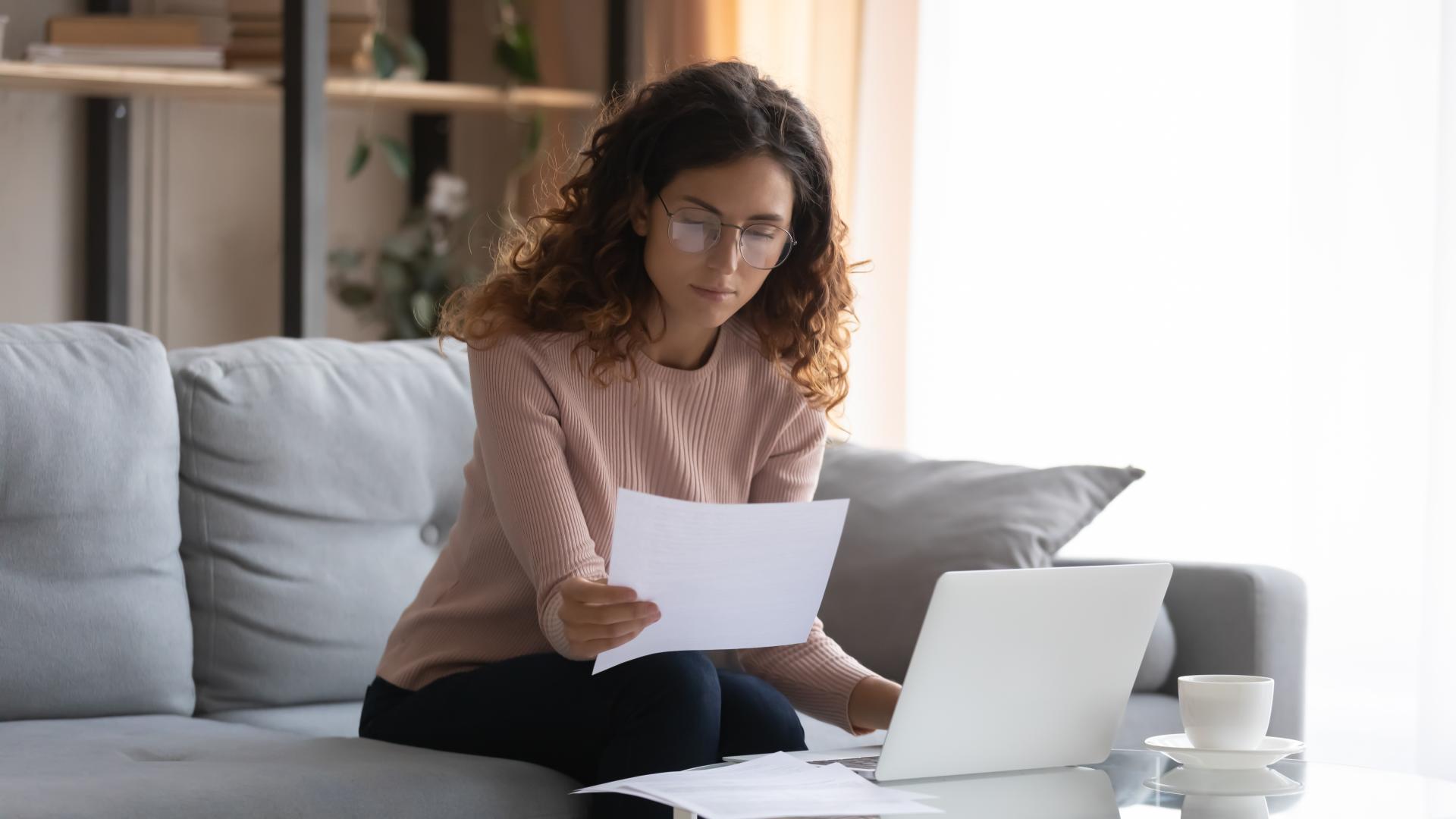 Woman sitting on couch in front of laptop computer holding paper